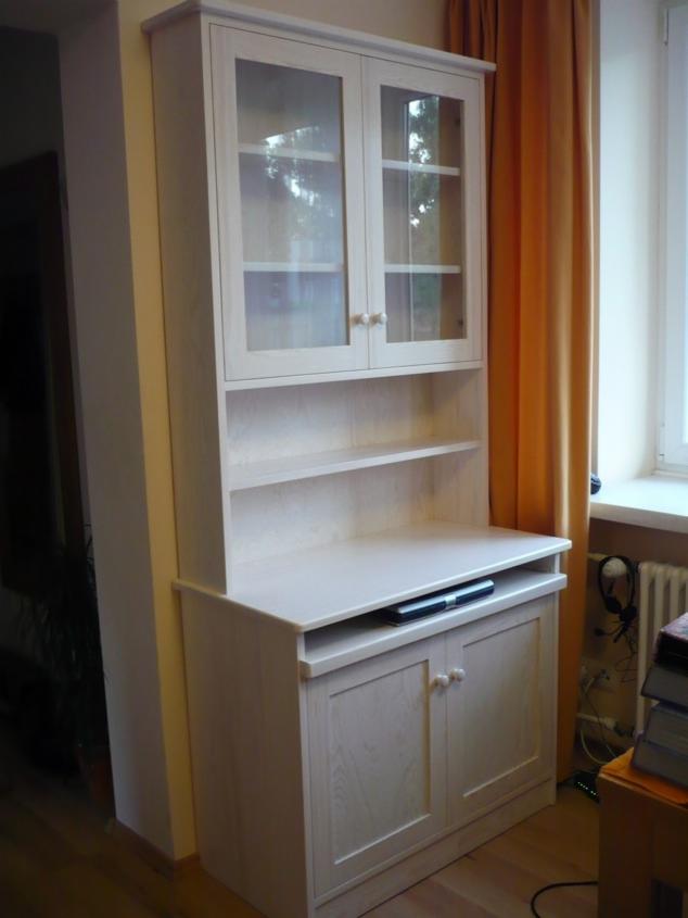 Jasna, drewniana witryna z przeszklonymi szafkami i otwartymi półkami, umieszczona przy oknie z pomarańczową zasłoną. Widoczne detale wykończenia i naturalne światło.