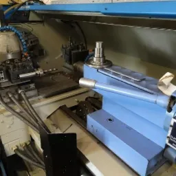 Precyzyjna tokarka CNC w trakcie obróbki metalu, widoczne narzędzia i wióry, oświetlenie warsztatowe, detale mechaniczne, manometr.