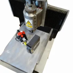 Frezer CNC z silnikiem i sterownikiem, widok z góry pod kątem. Metalowa konstrukcja, czarne osłony, żółta naklejka ostrzegawcza. Sprzęt do obróbki skrawaniem.