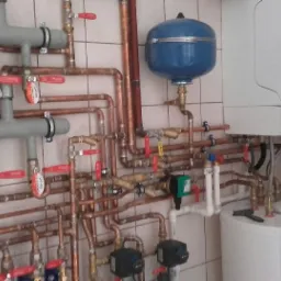 Kompleksowa instalacja centralnego ogrzewania z kotłem gazowym, zasobnikiem ciepłej wody i rozbudowaną siecią rur miedzianych z zaworami i manometrami na tle kafelkowanej ściany.
