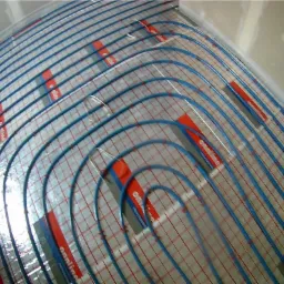 Rozłożona mata grzewcza podłogowa z niebieskimi rurkami na siatce z czerwonymi elementami mocującymi, na tle surowej ściany.