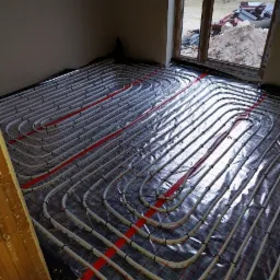 Białe rurki ogrzewania podłogowego ułożone na folii z czerwoną siatką, widok z góry w surowym wnętrzu w trakcie instalacji, okno z widokiem na plac budowy.