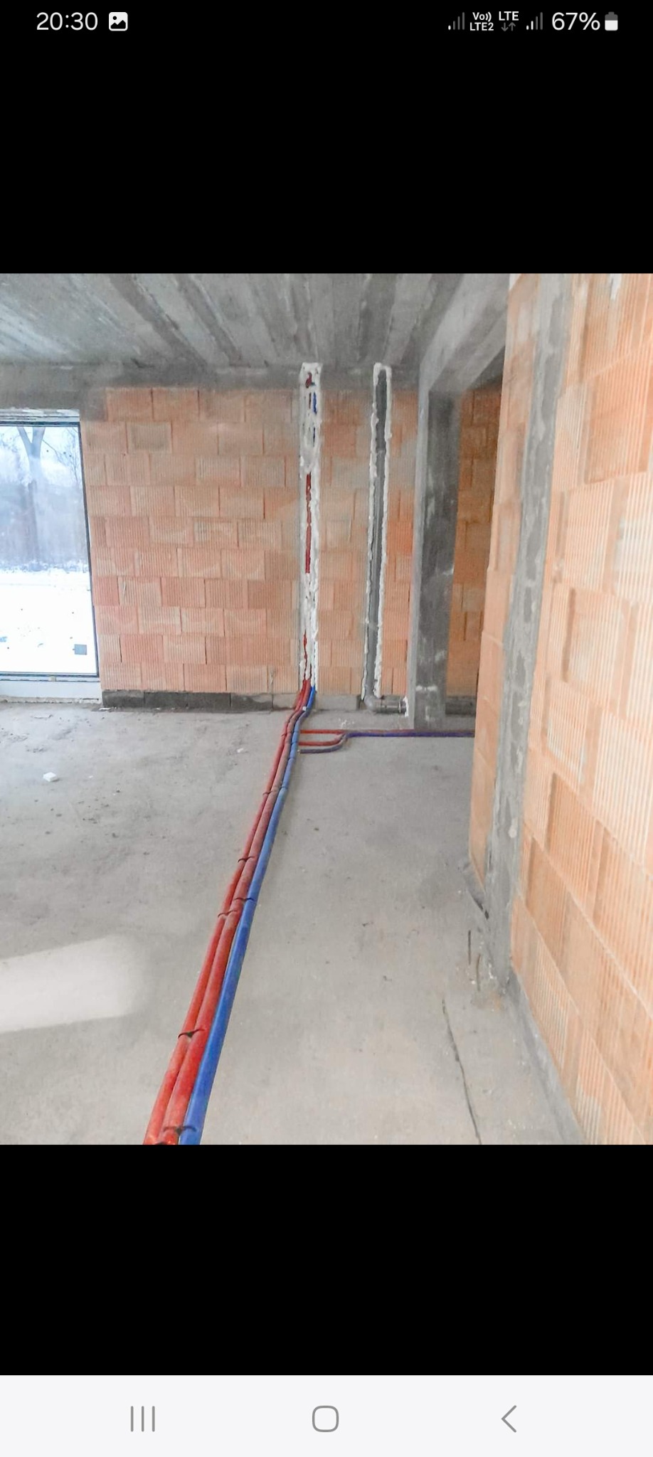 Wnętrze w stanie surowym z widocznymi, częściowo zamontowanymi instalacjami: czerwone i niebieskie rury prowadzone po podłodze i w bruzdach w ścianie z cegły.