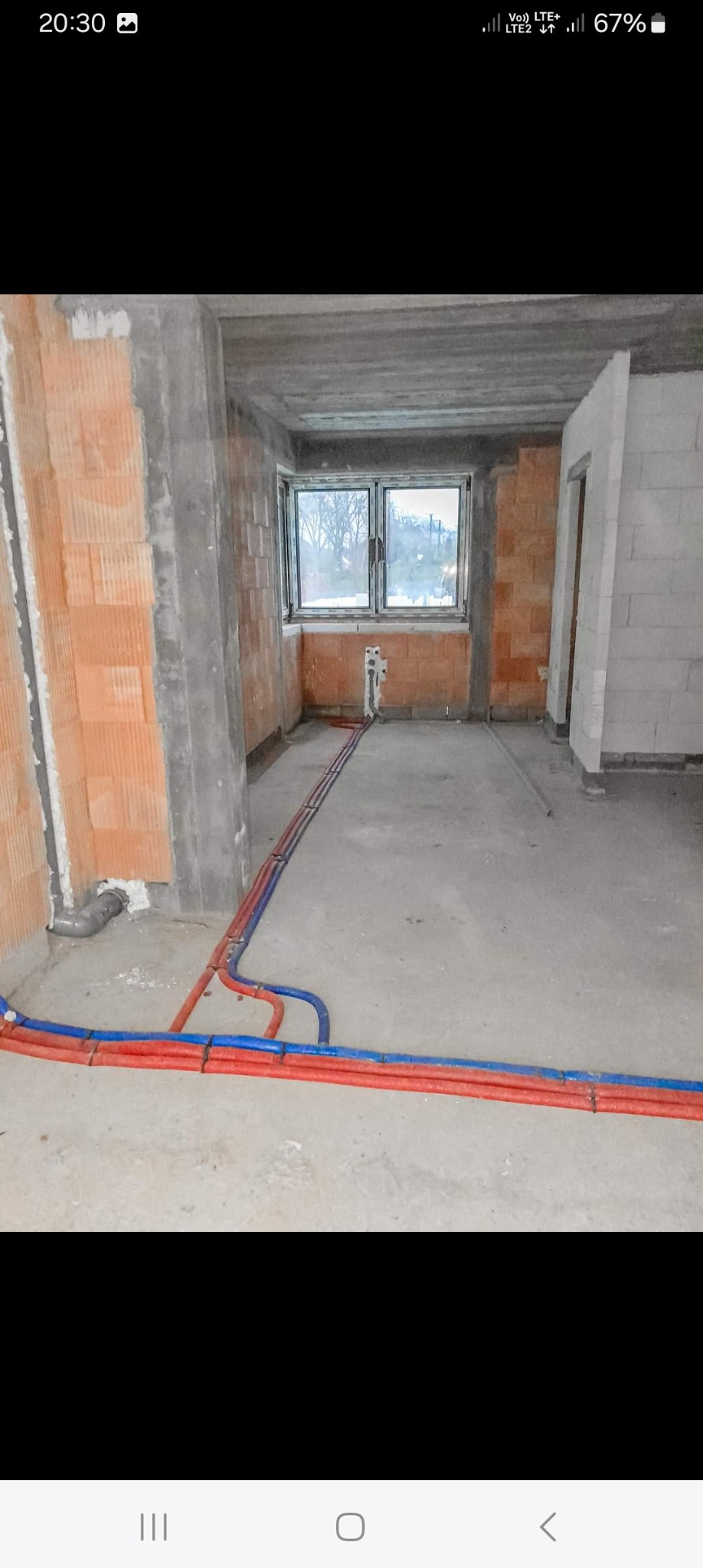 Wnętrze w trakcie budowy z widocznymi czerwono-niebieskimi rurami instalacji wodnej ułożonymi na surowej posadzce, ściany z cegły i betonu, okno w tle.
