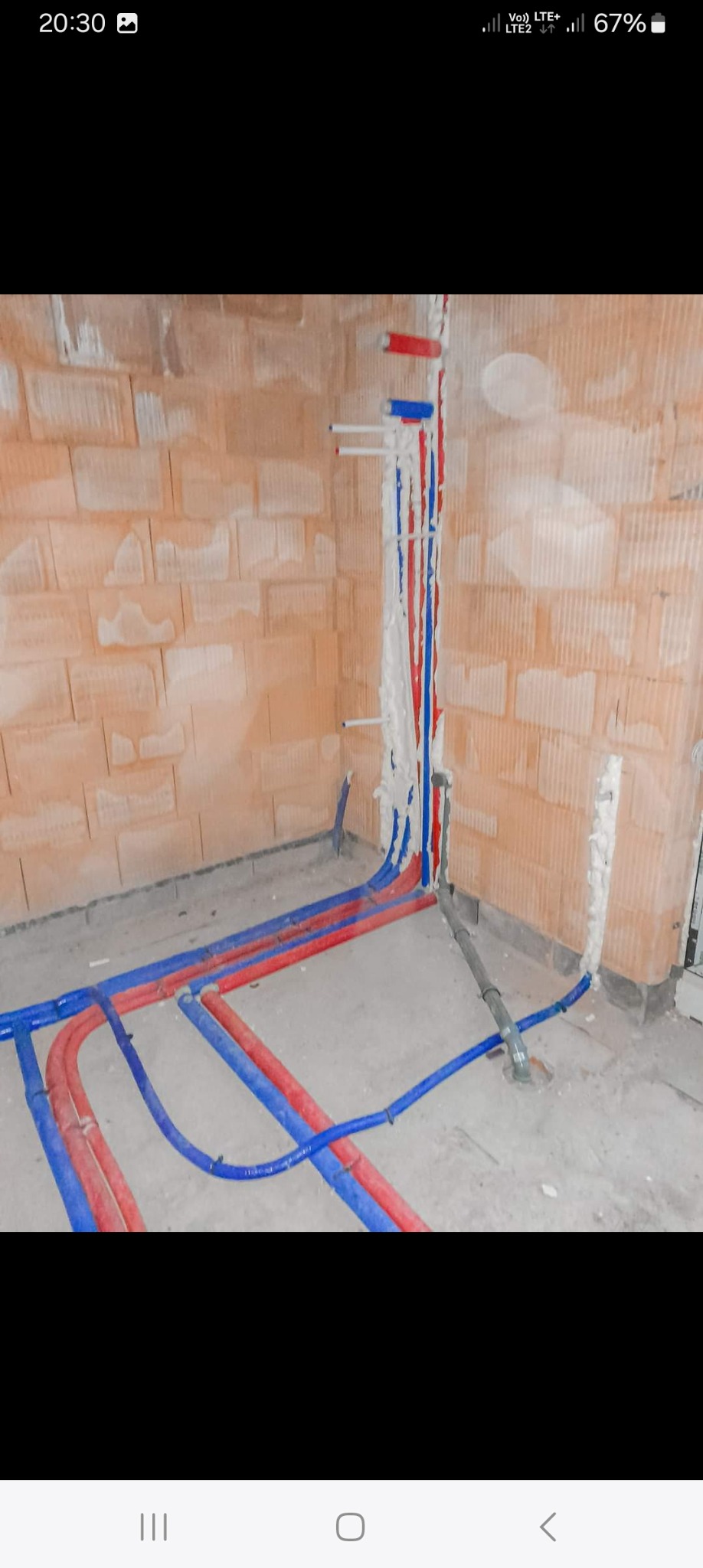 Surowe ściany z cegły z pionowo i poziomo ułożonymi rurami instalacji wodnej w kolorach czerwonym i niebieskim, częściowo zamaskowanymi pianką montażową.