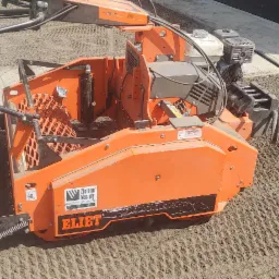 Pomarańczowa maszyna do przygotowywania podłoża pod trawnik, z widocznym wzorem w ziemi po jej przejeździe.