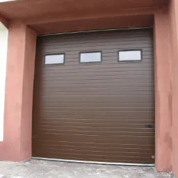 Brązowa segmentowa brama garażowa z trzema prostokątnymi okienkami u góry, zamontowana w brązowym portalu. Widoczny fragment szarej posadzki.