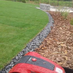 Czerwony robot koszący trawnik, zaparkowany w stacji dokującej, przy obrzeżu z kamieni oddzielającym trawnik od kory drzewnej, w tle zielony trawnik i drzewa.