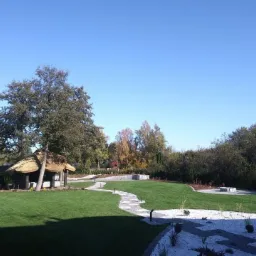 Widok na wypielęgnowany trawnik z altaną krytą strzechą, wijącą się ścieżką z kamienia i rabatami, w tle drzewa i fragment domu.