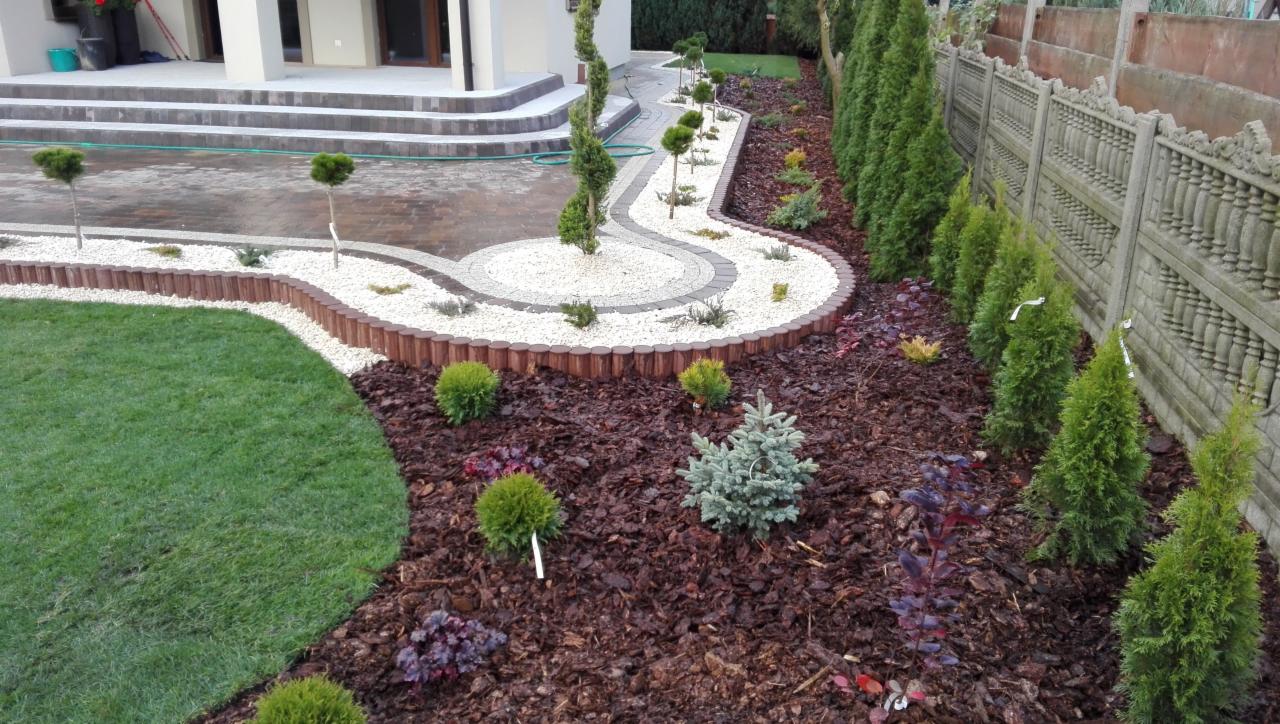 Ogród po metamorfozie: równo przystrzyżony trawnik graniczy z rabatą wysypaną korą, na której rosną iglaki, krzewy ozdobne i byliny. Ścieżka z białego grysu wijąca się między roślinami, obrzeżona...