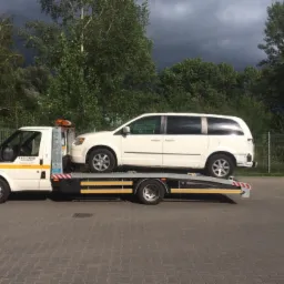 Biała laweta z załadowanym białym vanem na tle zieleni i pochmurnego nieba, widoczny napis '24H' na pojeździe.