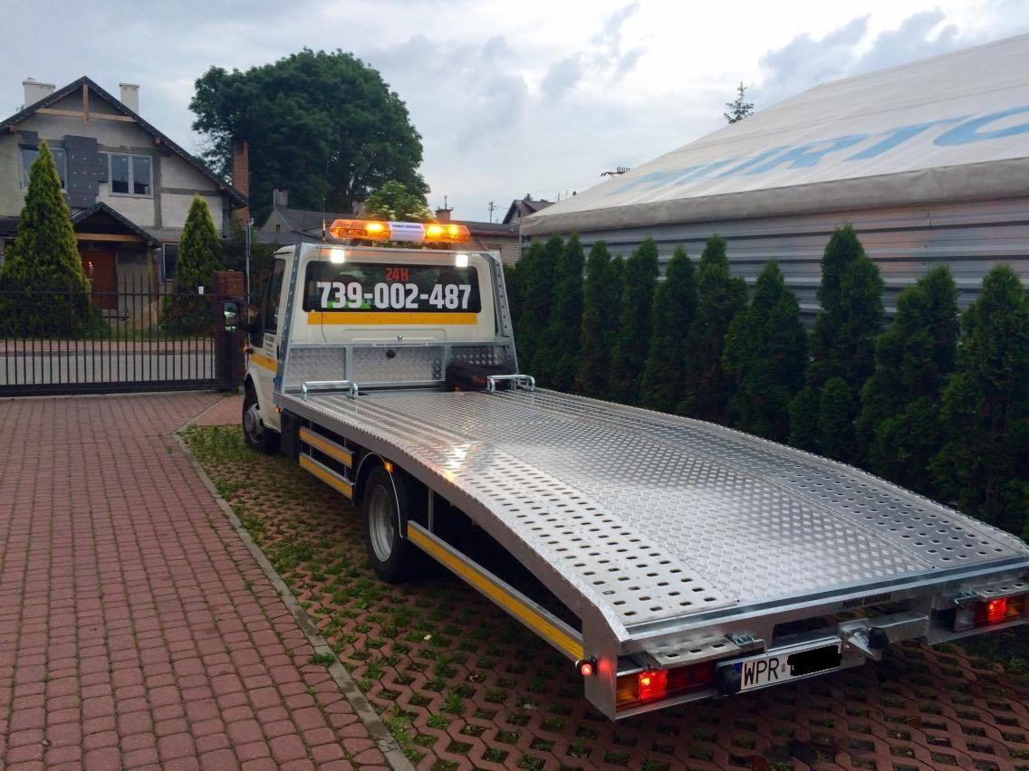 Pusta, aluminiowa platforma lawety na podwoziu samochodu ciężarowego, z włączonymi światłami ostrzegawczymi, zaparkowana na brukowanej posesji przed domem i wysokimi tujami.