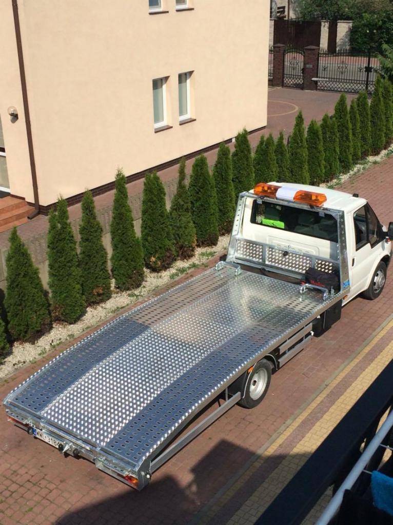 Biała laweta z aluminiową platformą w tle domu z tujami, widok z góry.