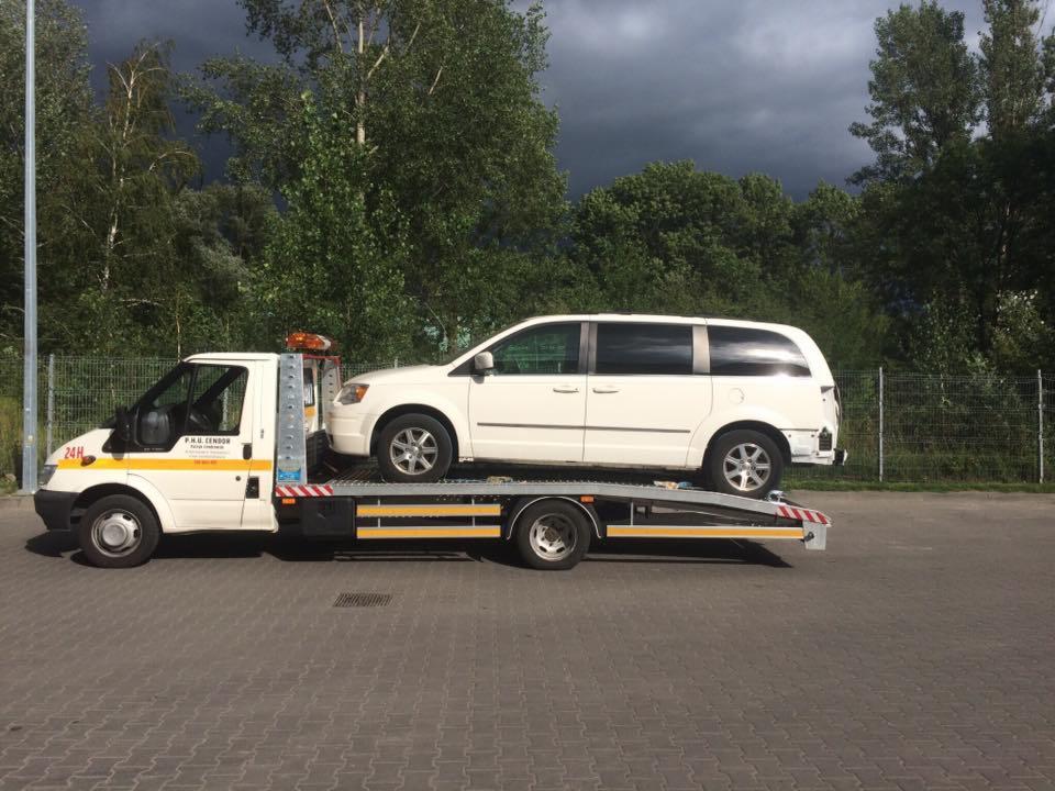 Biała laweta z załadowanym białym vanem na tle zieleni i pochmurnego nieba, widoczny napis '24H' na pojeździe.