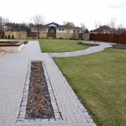 JKB Consulting.eu Jacek Kozica - Ułożona z kostki granitowej ścieżka z centralnym pasem kory prowadząca przez trawnik w ogrodzie, z widocznym tarasem i ogrodzeniem w tle.
