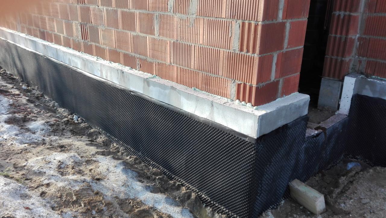 Ceglany mur w trakcie budowy z widoczną hydroizolacją fundamentów w postaci czarnej folii kubełkowej. Świeżo wylany betonowy fundament. Widoczny fragment terenu budowy.