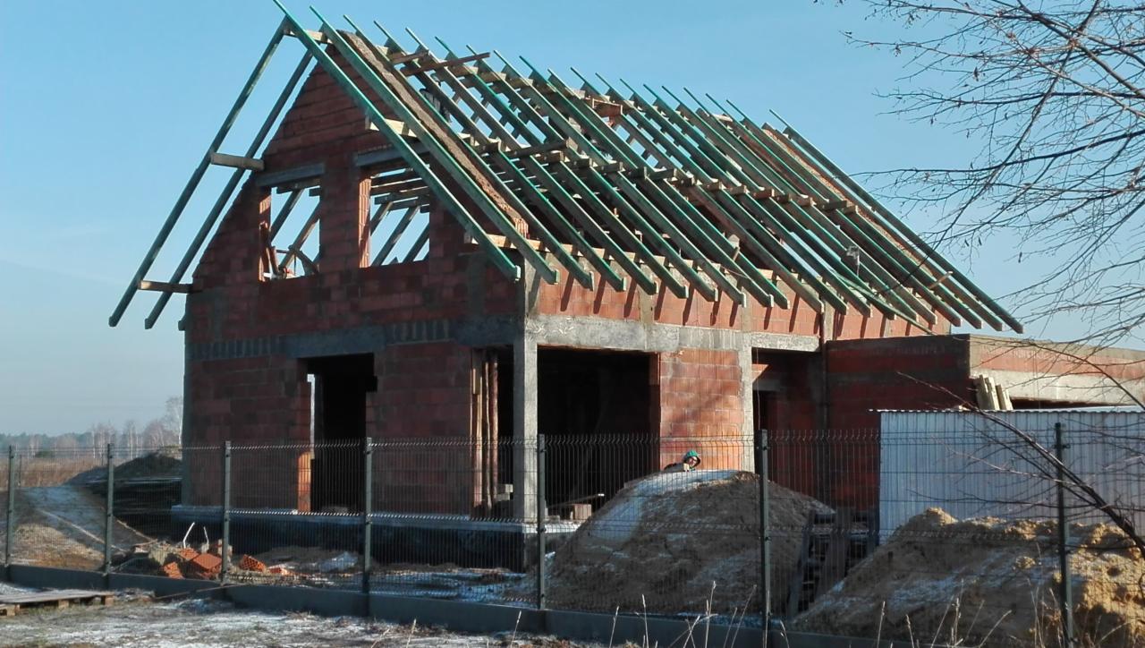 Surowy stan domu jednorodzinnego w trakcie budowy: czerwona cegła, drewniana konstrukcja dachu, ogrodzenie z siatki, sterta piasku, zimowy krajobraz w tle.