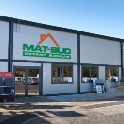 Fasada budynku z dużym napisem 'MAT-BUD MATERIAŁY BUDOWLANE', ekspozycja dachówek przed wejściem, zaplecze z ułożonymi paletami materiałów budowlanych, widok z zewnątrz.