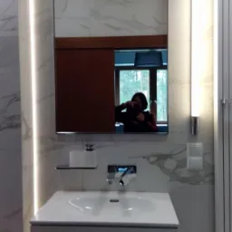 Wykończona łazienka z umywalką, lustrem i oświetleniem LED na marmurowych płytkach. W lustrze odbija się osoba robiąca zdjęcie, a za nią widok na zewnątrz.