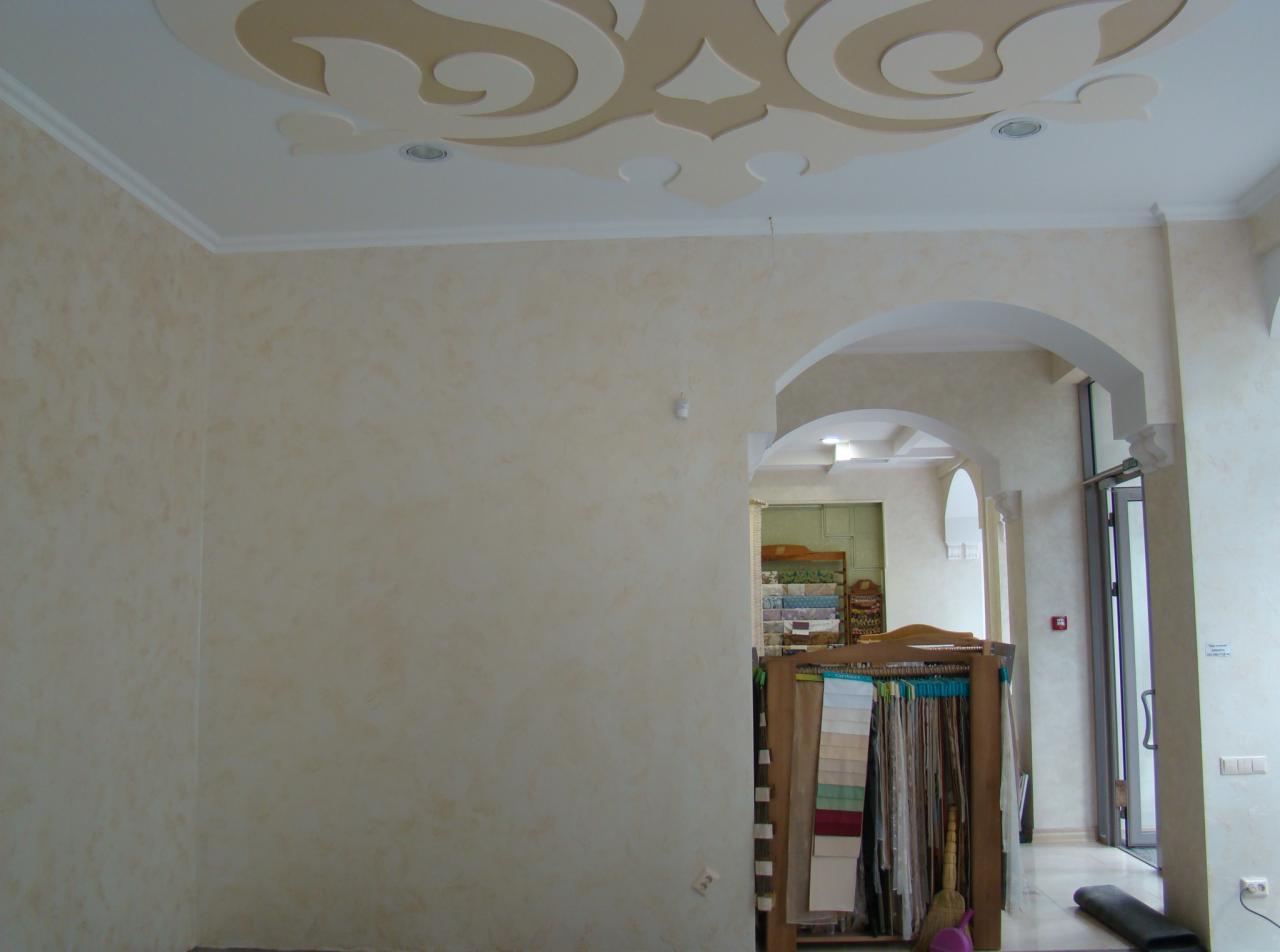 Jasne ściany z efektem dekoracyjnym i zdobiony sufit w odcieniach beżu. Widoczne łuki architektoniczne i fragment ekspozycji tkanin w głębi pomieszczenia.