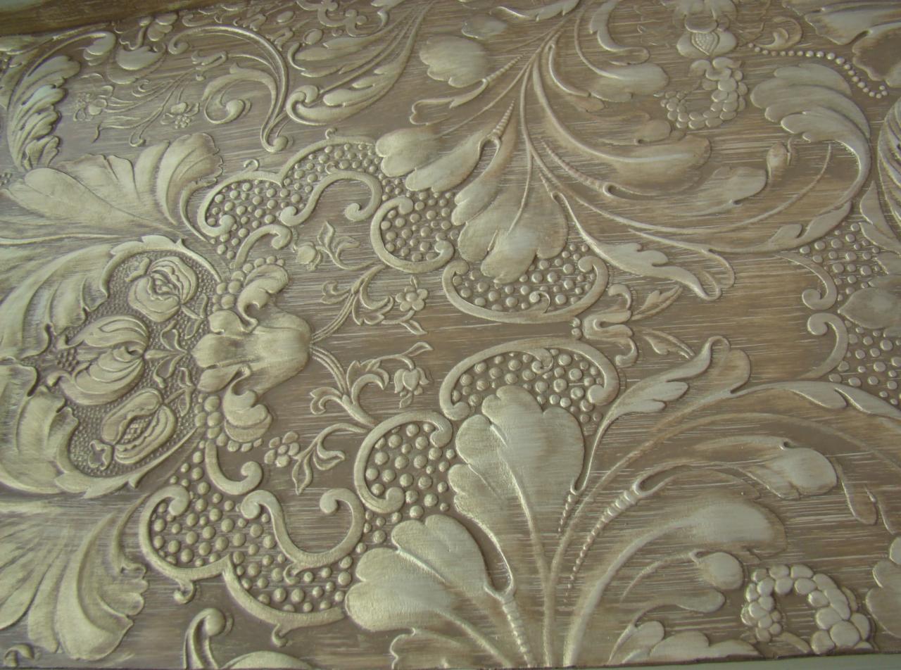Dekoracyjna płaskorzeźba na drewnianej powierzchni w odcieniach beżu i złota, przedstawiająca motywy roślinne: liście, kwiaty i winogrona. Delikatne światłocienie podkreślają detale.