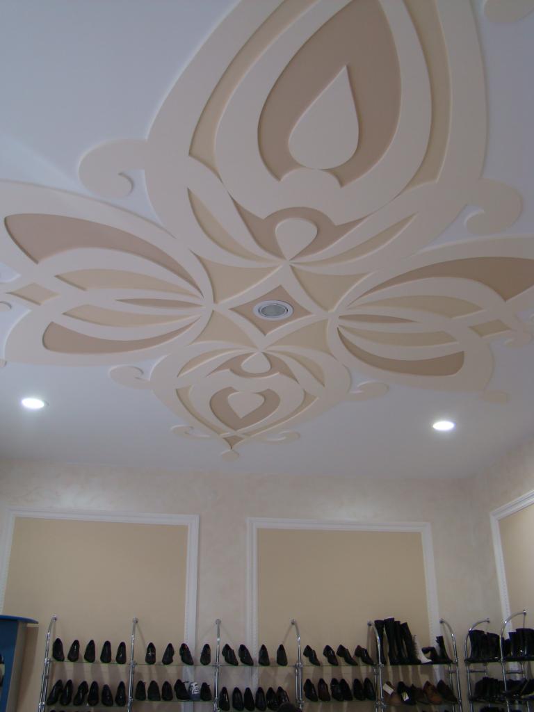 Dekoracyjny sufit z gładzią w kremowych odcieniach, z centralnym wzorem kwiatowym i oświetleniem punktowym. Widok z dołu, w tle ściana z ramami.