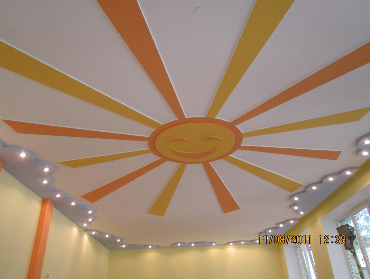 Biały sufit z ręcznie malowanym, uśmiechniętym słońcem w odcieniach żółci i pomarańczy, oraz dekoracyjnymi obniżeniami w kształcie chmur z oświetleniem punktowym.