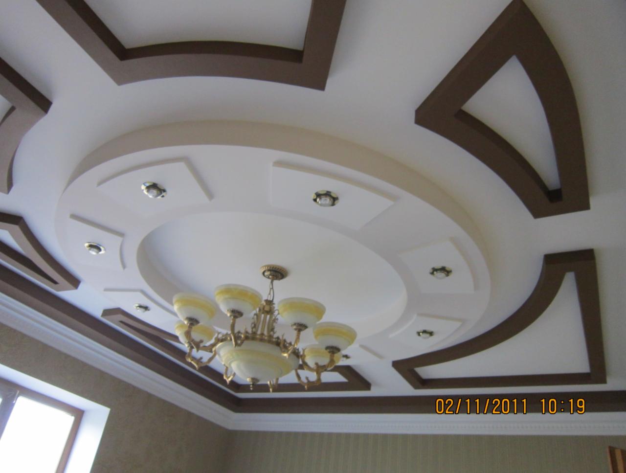 Dekoracyjny sufit podwieszany z oświetleniem punktowym i żyrandolem. Brązowe elementy zdobnicze tworzą geometryczny wzór na białym tle. Ujęcie z dołu.