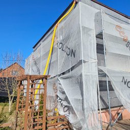 Eco Solution Rybnik - Dom w trakcie prac ociepleniowych, osłonięty siatką z nadrukiem, widoczne rusztowania i żółty przewód wentylacyjny. W tle drugi dom i drzewa.