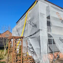 Dom w trakcie prac ociepleniowych, osłonięty siatką z nadrukiem, widoczne rusztowania i ż&oacute;łty przew&oacute;d wentylacyjny. W tle drugi dom i drzewa.