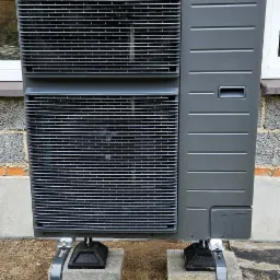 Szara pompa ciepła Panasonic Aquarea zamontowana na betonowych bloczkach z metalową konstrukcją wsporczą, widoczna na tle ściany z szarej cegły.