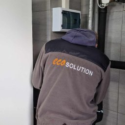 Eco Solution - Instalator w bluzie z logo 'eco SOLUTION' montuje rury instalacji grzewczej w pomieszczeniu wyłożonym szarą płytką, widoczna skrzynka elektryczna na ścianie.