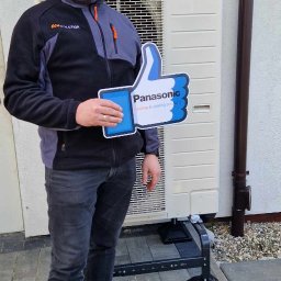 Eco Solution - Mężczyzna trzyma kciuk w górę z logo Panasonic przed zainstalowaną jednostką zewnętrzną pompy ciepła na tle jasnej ściany budynku.