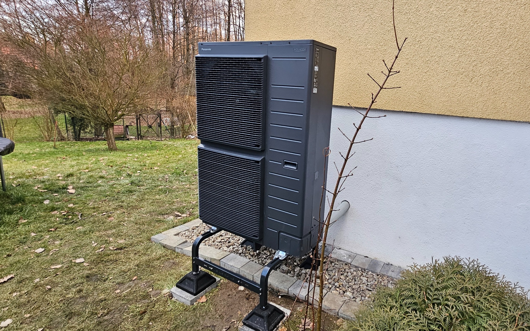 Szara jednostka zewnętrzna z logo Panasonic Aquarea, zamontowana na czarnej konstrukcji wsporczej, na tle ściany budynku i trawnika z drzewami.