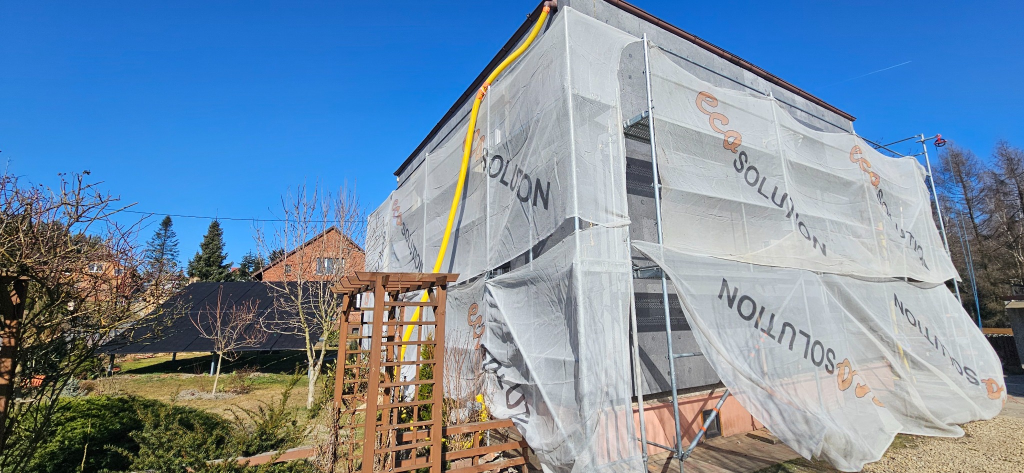 Dom w trakcie prac ociepleniowych, osłonięty siatką z nadrukiem, widoczne rusztowania i żółty przewód wentylacyjny. W tle drugi dom i drzewa.