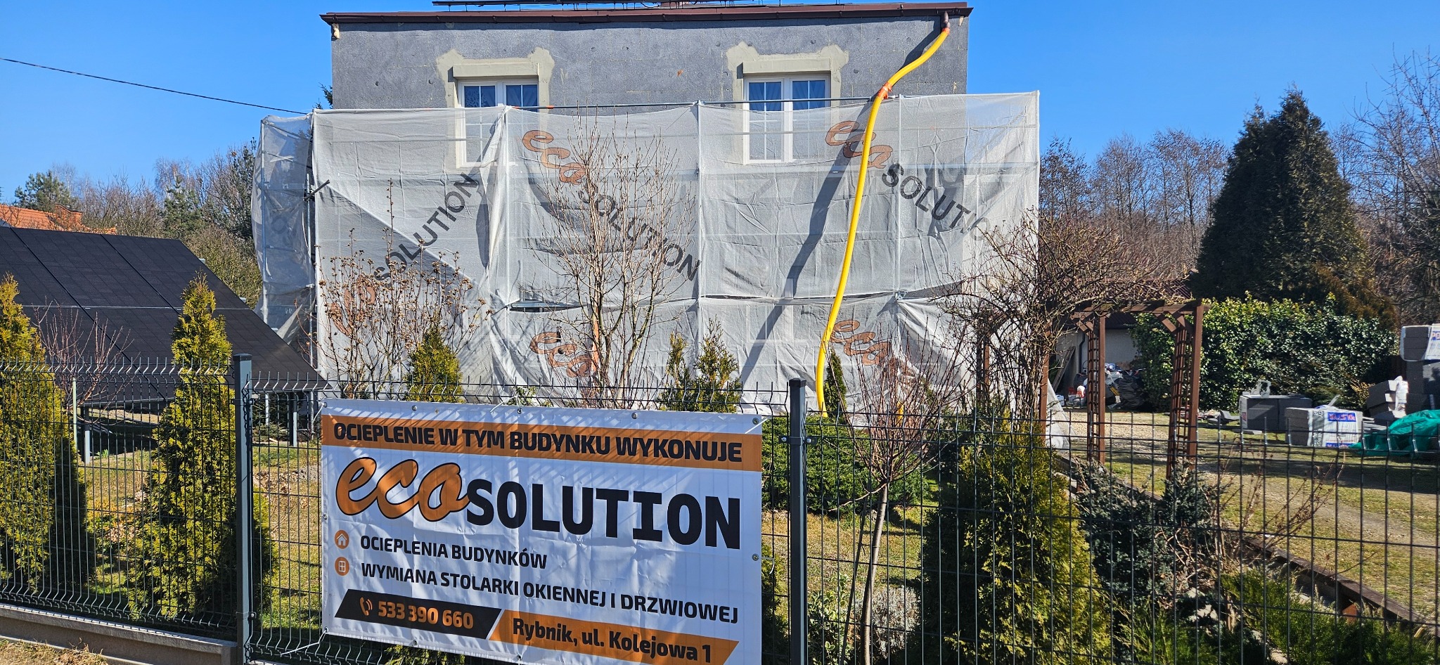 Budynek w trakcie ocieplania elewacji, osłonięty siatką z nadrukowanym logo firmy EcoSolution, na pierwszym planie baner reklamowy tej samej firmy.