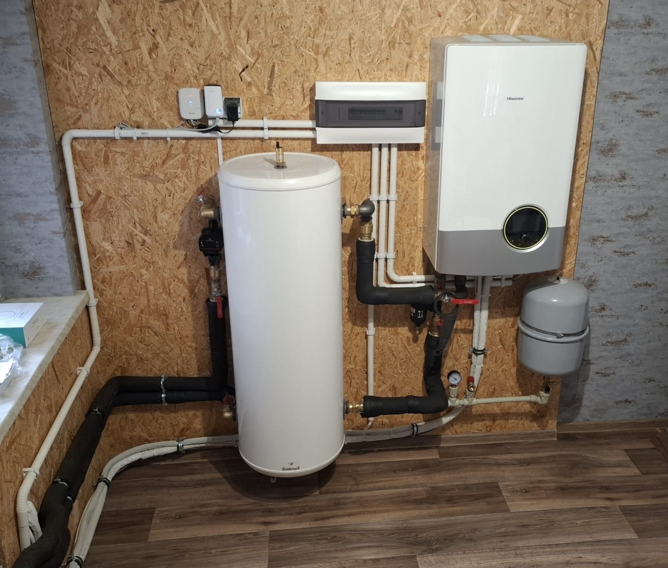 Biały zasobnik ciepłej wody użytkowej Galmet i wiszący, szary kocioł Hisense z podłączeniami rur w kotłowni wykończonej płytami OSB.
