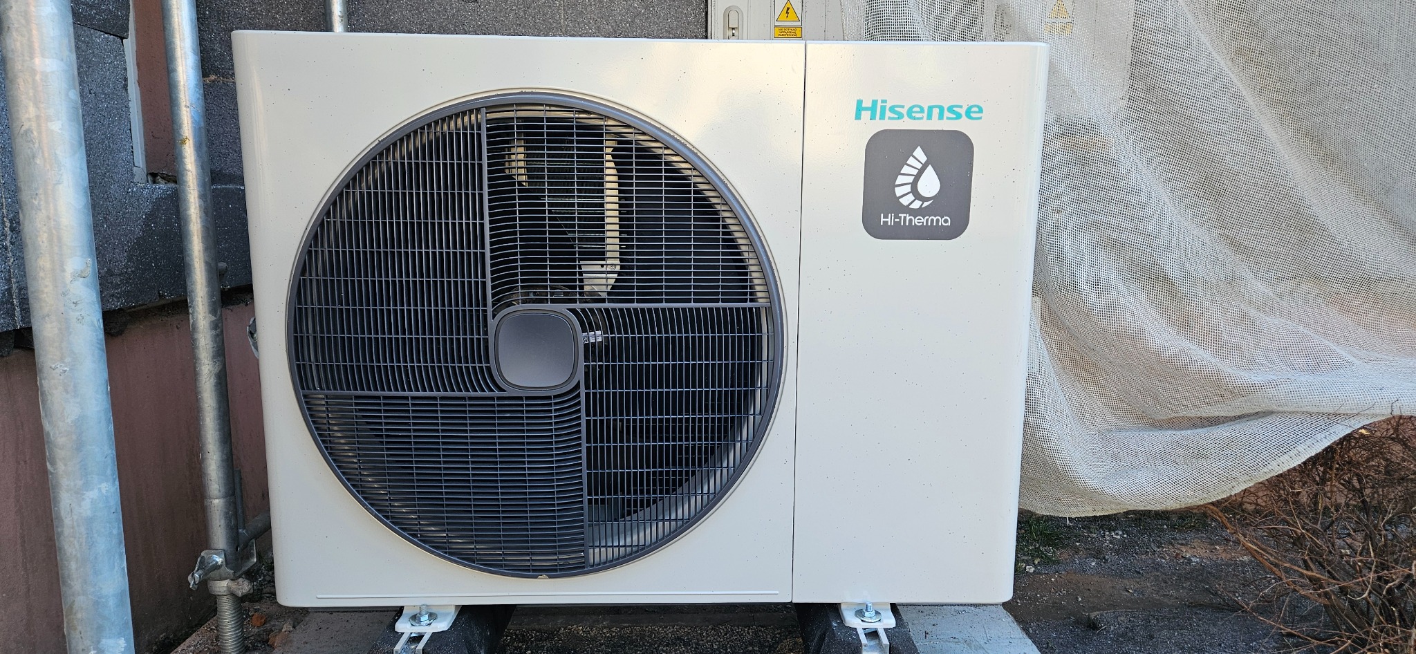 Zewnętrzna jednostka pompy ciepła Hisense Hi-Therma zamontowana na szarej podstawie, obok metalowych rurek rusztowania i osłonięta płótnem ochronnym.