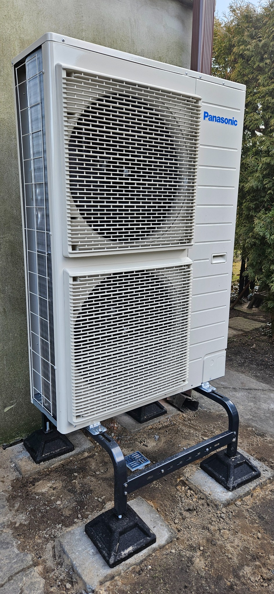 Zewnętrzna jednostka dwusekcyjnej, jasnej pompy ciepła marki Panasonic, zamontowana na czarnej, metalowej podstawie, widoczna na tle betonowej ściany i drzew.