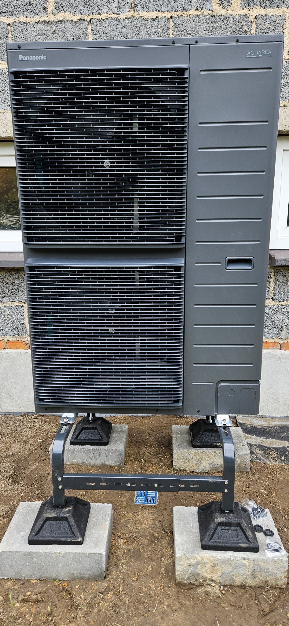 Szara pompa ciepła Panasonic Aquarea zamontowana na betonowych bloczkach z metalową konstrukcją wsporczą, widoczna na tle ściany z szarej cegły.