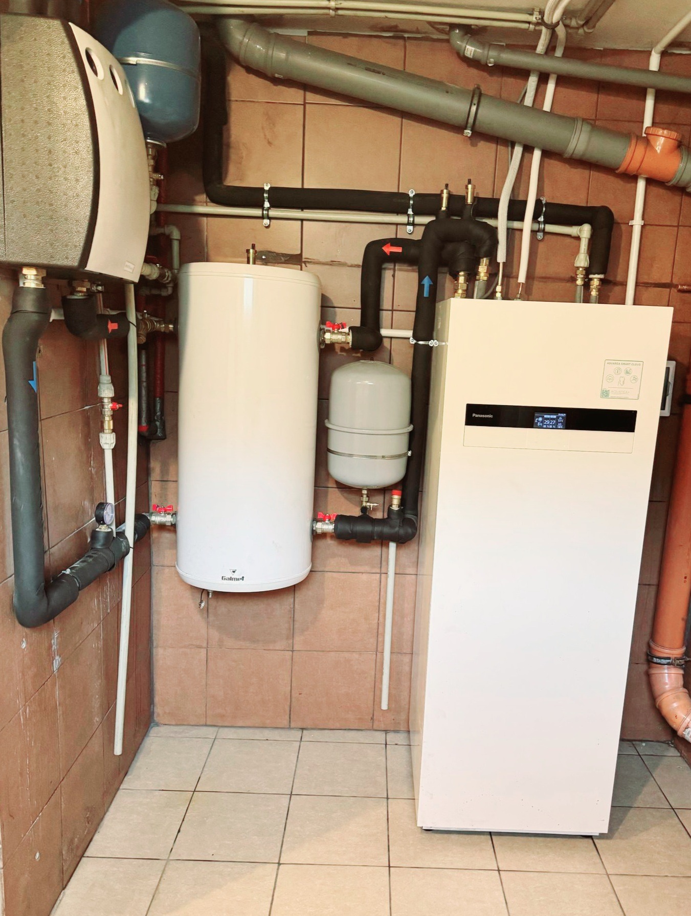 Instalacja grzewcza w kotłowni: biały zbiornik Galmet, szary zbiornik przeponowy i panel sterowania Panasonic Aquarea Smart Cloud, liczne rury i zawory, brązowe płytki na ścianach i podłodze.