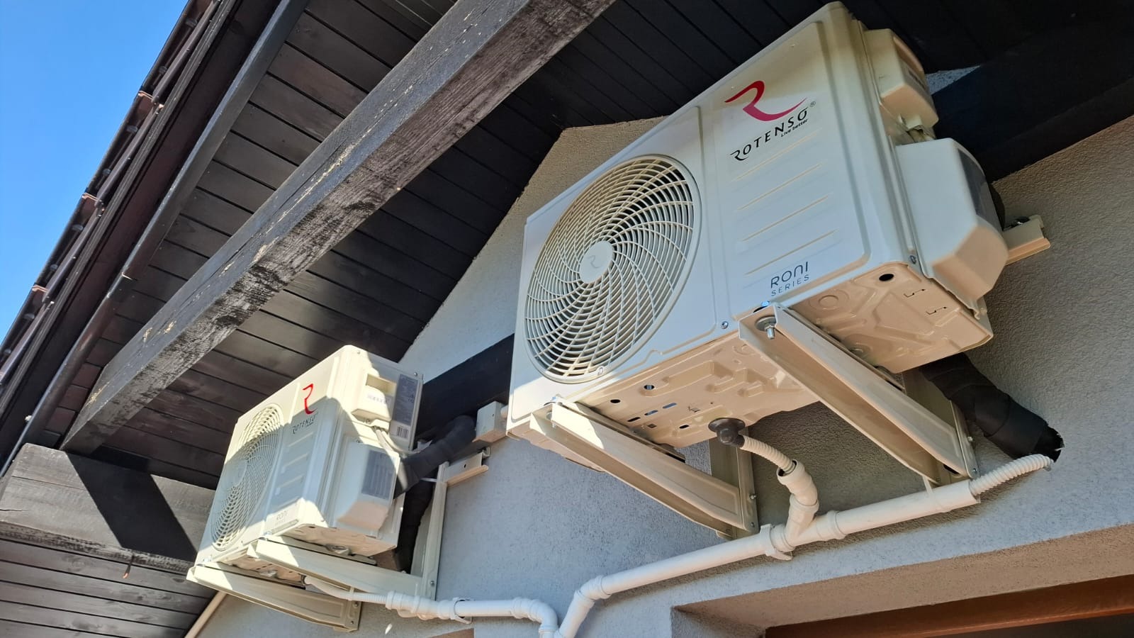 Dwie jednostki zewnętrzne klimatyzacji Rotenso Roni Series zamontowane na elewacji domu z widocznymi elementami instalacji i drewnianą podbitką dachu.