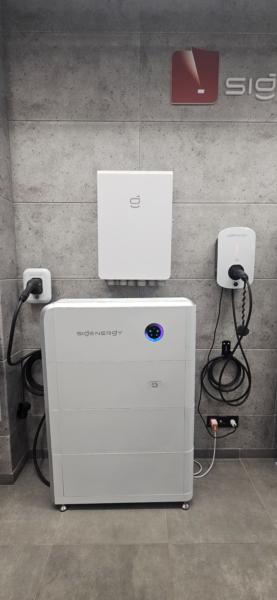 Biały magazyn energii z podłączonymi kablami na tle szarej ściany z logo firmy. Widoczne gniazdka i elementy instalacji elektrycznej. Minimalistyczny design.