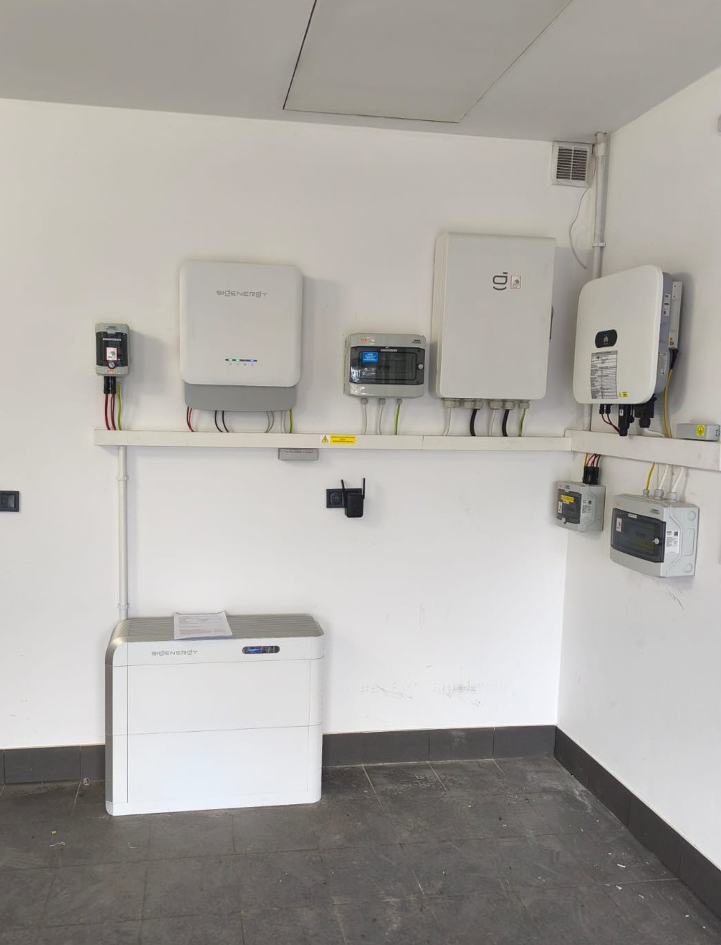 Białe urządzenia elektryczne, w tym magazyn energii, falowniki i skrzynki, zamontowane na ścianie w pomieszczeniu. Widoczne przewody i elementy instalacji.