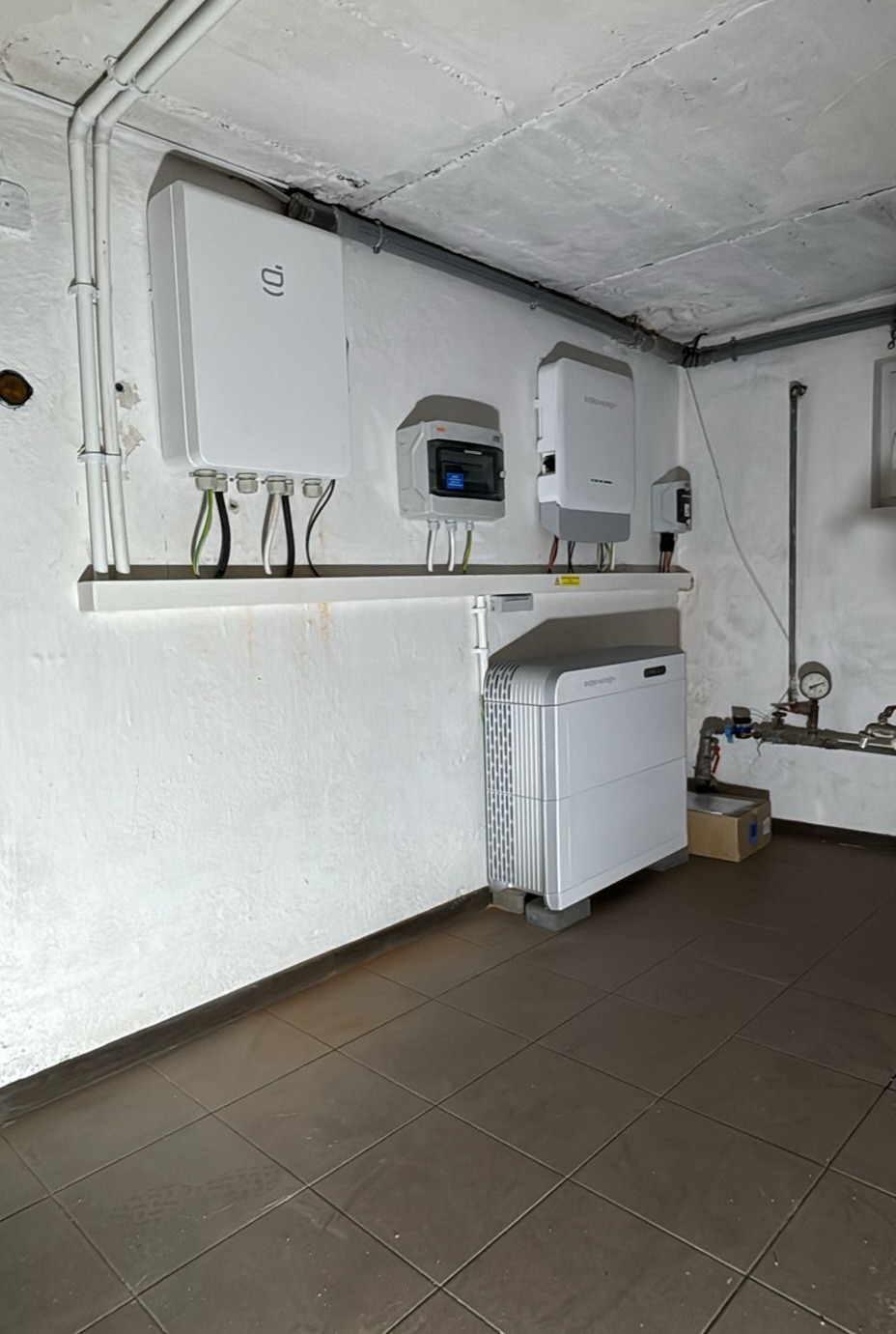 Biała instalacja z przewodami na ścianie w piwnicy. Widoczne urządzenia elektryczne, rury i brązowe płytki na podłodze. Instalacja w pomieszczeniu o surowym wykończeniu.