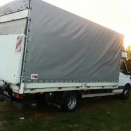 Biała ciężarówka Iveco z szarą plandeką na trawie, widok z tyłu. Budynek mieszkalny i drzewa w tle. Tablica rejestracyjna widoczna.