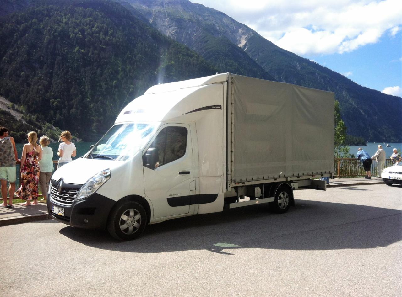 Biała furgonetka Renault Master z plandeką na tle górskiego krajobrazu z jeziorem i turystami, na parkingu.