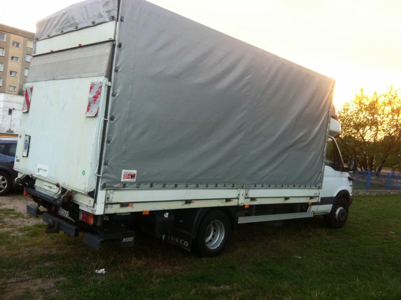 Biała ciężarówka Iveco z szarą plandeką na trawie, widok z tyłu. Budynek mieszkalny i drzewa w tle. Tablica rejestracyjna widoczna.