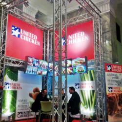 Stoisko United Chicken na targach: czerwone banery z logo, metalowa konstrukcja, monitory z menu, osoby w środku. Widok z boku, oświetlenie targowe.