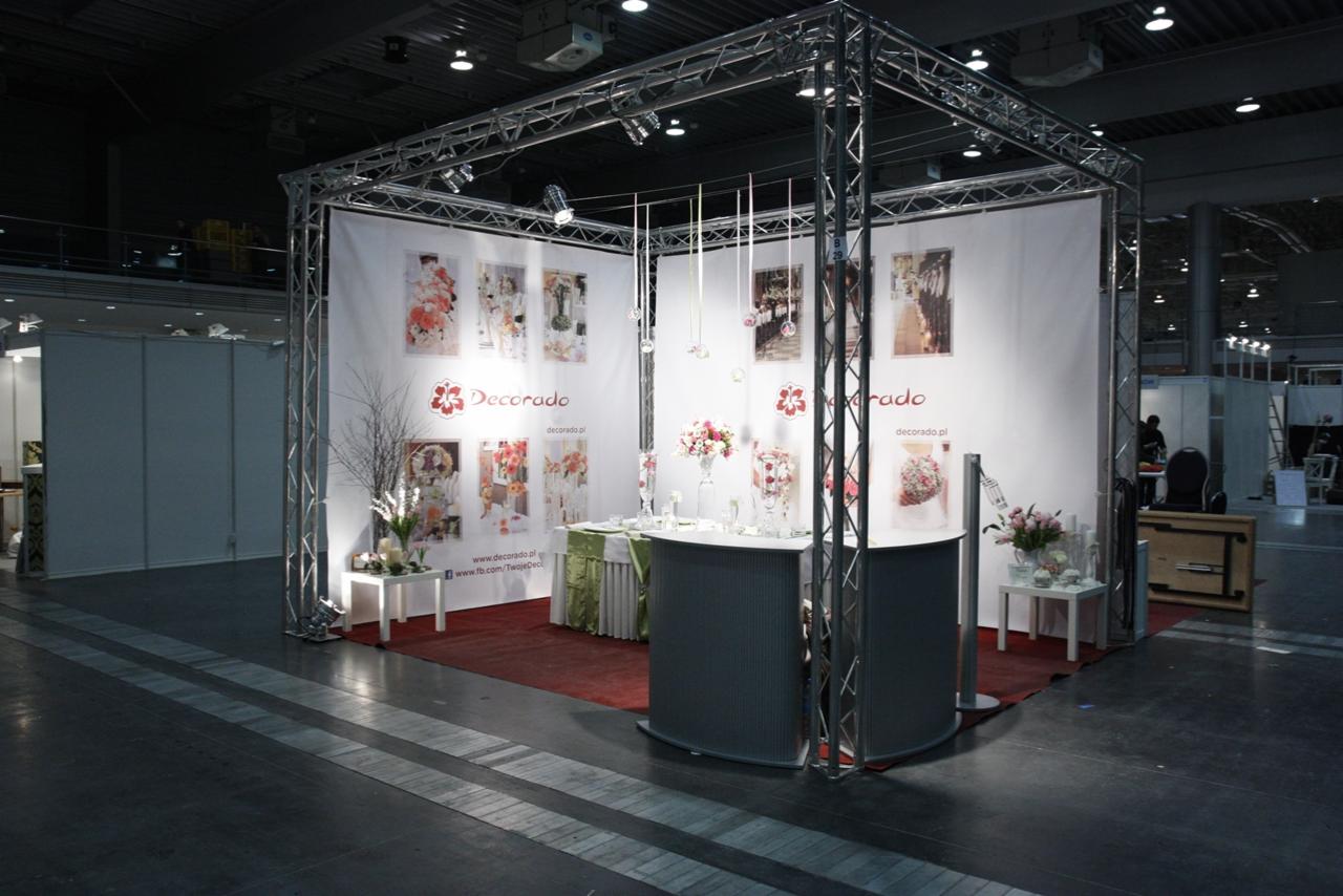 Stoisko targowe firmy Decorado z ekspozycją dekoracji kwiatowych i aranżacji ślubnych. Konstrukcja metalowa, białe ściany z grafikami, kwiaty w wazonach, oświetlenie punktowe.